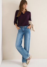 Mørkeblå bluse med V-udskæring og oprettede ærmer, parret med lyseblå bredbenede jeans. Hvide sneakers med bordeaux farvede detaljer fuldender looket.