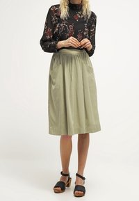 Blusa nera a fiori con maniche lunghe, abbinata a una gonna alta di colore verde oliva chiaro con tasche laterali e finitura testurizzata. Sandali neri.