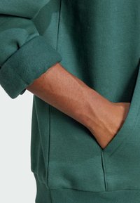 Fazzoletto verde foresta in tessuto, con polsino ripiegato, presenta una definizione della tasca laterale e una texture liscia. Sezione di cucitura sottile visibile.