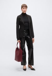 Μαύρο σακάκι πάνω από μαύρο ζιβάγκο, συνδυασμένο με μαύρο παντελόνι υψηλής λάμψης. Κρατάει μια μπορντό τσάντα tote. Μαύρες μυτερές γόβες ολοκληρώνουν την εμφάνιση.