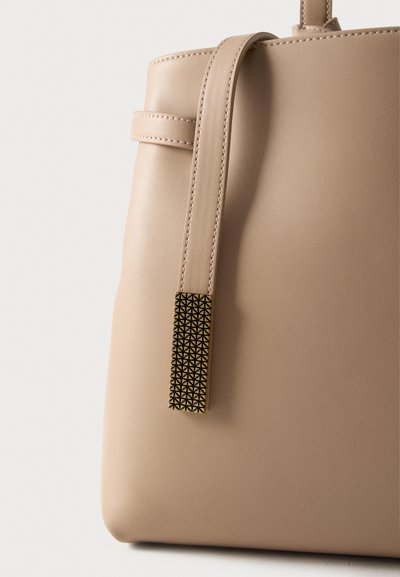 Μπεζ δερμάτινη τσάντα με ομαλή υφή, που διαθέτει μακρύ λουρί και διακοσμητικό εξάρτημα σε χρυσαφί απόχρωση με γεωμετρικό μοτίβο.