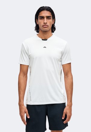 Homme aux cheveux foncés portant une chemise blanche unie à manches courtes et un short noir, debout devant un fond clair.