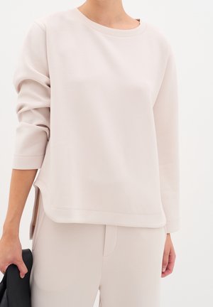 Femme portant un haut à manches longues beige clair avec des fentes latérales et un pantalon assorti, tenant un objet noir plié dans sa main gauche.