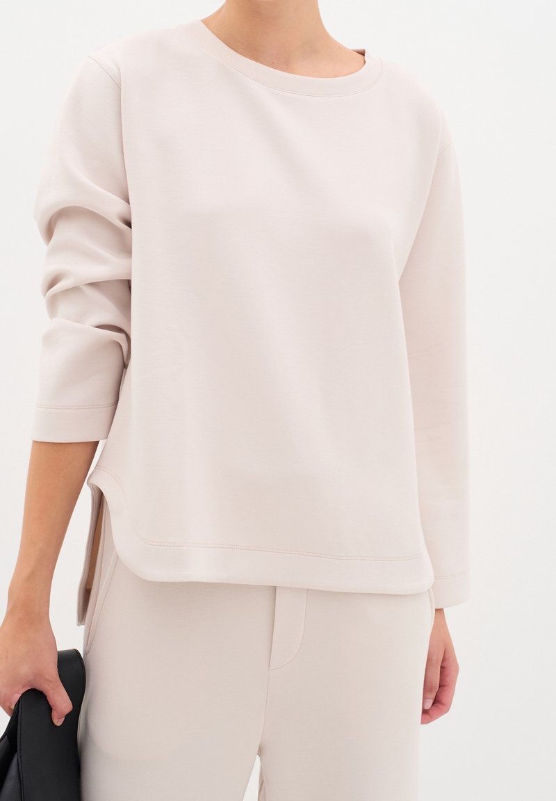 Femme portant un haut à manches longues beige clair avec des fentes latérales et un pantalon assorti, tenant un objet noir plié dans sa main gauche.