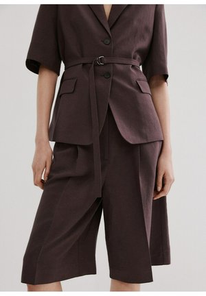 Mujer vistiendo un blazer de manga corta marrón oscuro con cinturón y pantalones cortos de pierna ancha a la altura de la rodilla a juego, sobre un fondo sencillo.