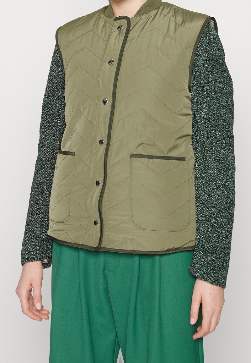 Gilet matelassé vert olive avec un motif en chevrons, col rond, boutons pression et deux poches avant, superposé sur une chemise à manches longues texturée verte.