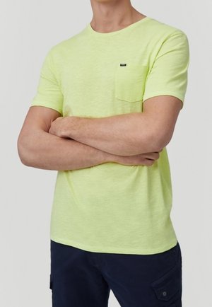 Homme les bras croisés portant un t-shirt jaune clair à manches courtes avec une poche sur la poitrine et un pantalon bleu foncé sur fond clair.