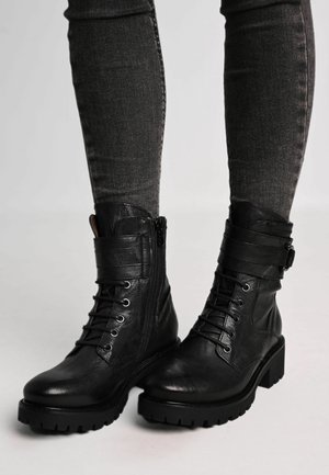 Bottes mi-mollet en cuir noir avec bout arrondi, fermeture à lacets sur le devant, fermeture éclair latérale, et semelle épaisse en caoutchouc. Comprennent des brides près du haut.