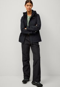 Giacca impermeabile nera con cappuccio, indossata sopra una camicia verde. Abbinata a pantaloni impermeabili neri e scarpe nere robuste.
