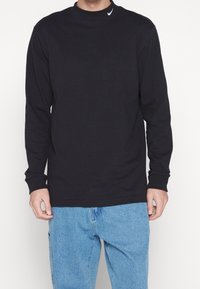 Černé tričko s dlouhým rukávem z bavlny, s kulatým výstřihem a malým bílým logem Nike na límci. Kombinováno s světle modrými džínovými kalhotami.