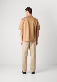Mann som står med ryggen mot kameraet, iført en beige kortermet skjorte, beige bukser og svarte sko mot en lys, ensfarget bakgrunn.