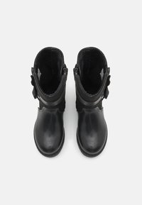 Mustat nilkkurit kiiltävällä pinnalla, tekstuurilla varustetut varret, sivu vetoketjukiinnitys ja koristeelliset nauhat kummallakin puolella. Lämpimällä fleecevuorilla varustettu sisäosa.