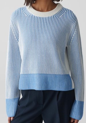 Femme portant un pull en maille à rayures verticales bleu et blanc avec poignets et ourlet bleu uni, associé à un pantalon foncé sur un fond uni.
