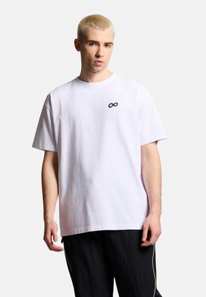 Młody mężczyzna z krótkimi blond włosami, ubrany w białą, prostą koszulkę z małym czarnym symbolem nieskończoności na piersi.