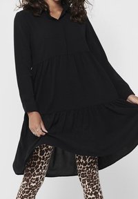 Abito nero a maniche lunghe con colletto, caratterizzato da un design a strati e una silhouette fluida. Indossato sopra leggings con stampa leopardata.