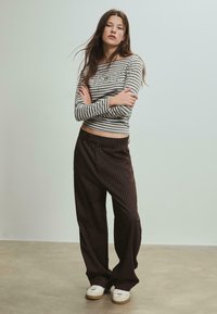 Prúžkované dlhé rukávy v sivej a bielej farbe, spojené s voľnými, tmavo hnedými prúžkovanými nohavicami. Model má na sebe biele tenisky. Minimalistické pozadie.