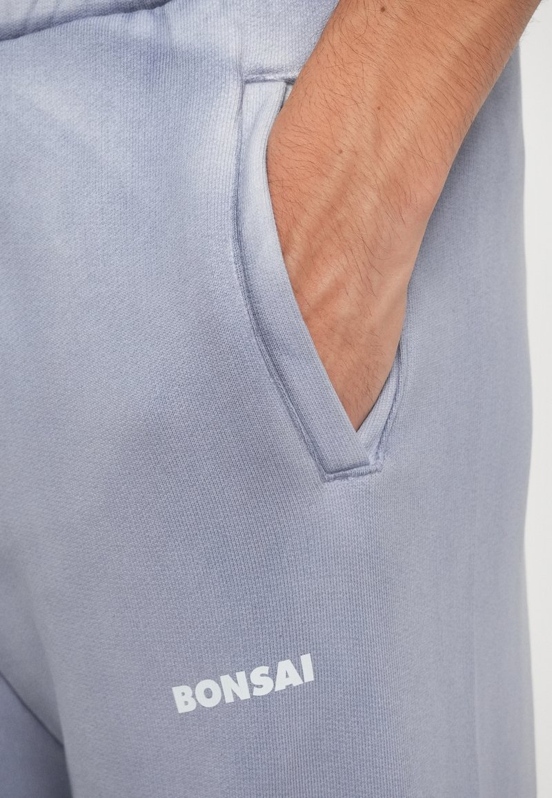 Pantaloni jogger grigi realizzati in tessuto morbido, con tasca laterale e logo "BONSAI" in testo bianco. Texture liscia con design minimale.