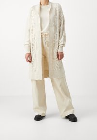 Osoba v krémovém pleteném kardiganu dlouhém přes béžové široké kalhoty a světlém topu, doplněná černými botami, stojící na bílém pozadí.