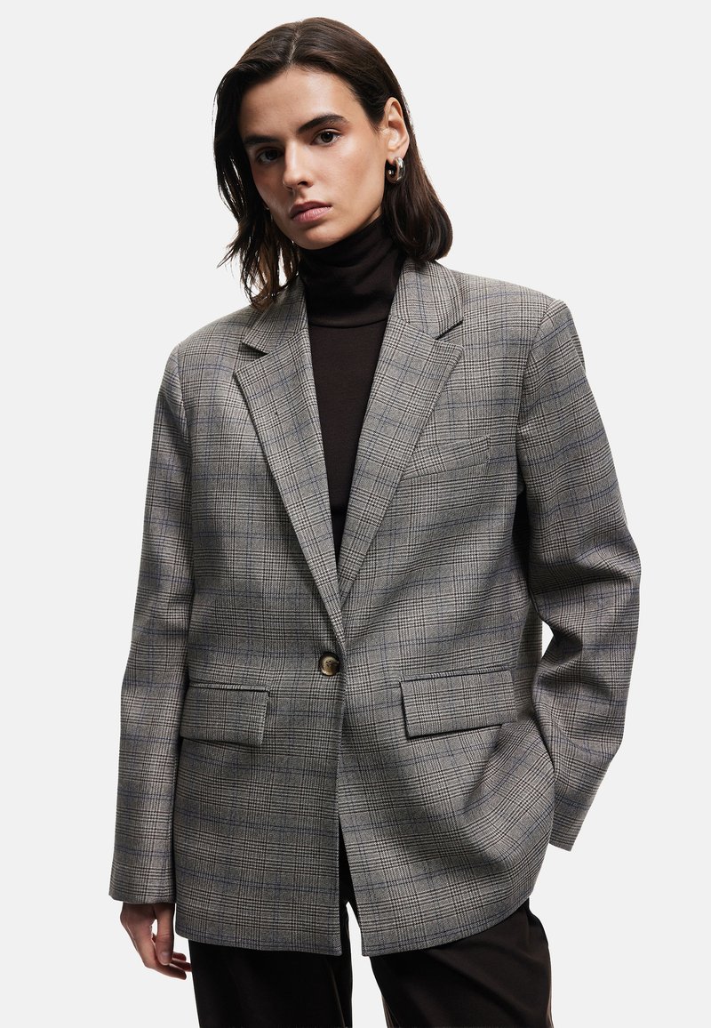 Grå ternet blazer med en struktureret pasform, reverskrave, to forlommer og en enkelt knaplukning. Båret over en sort turtleneck.