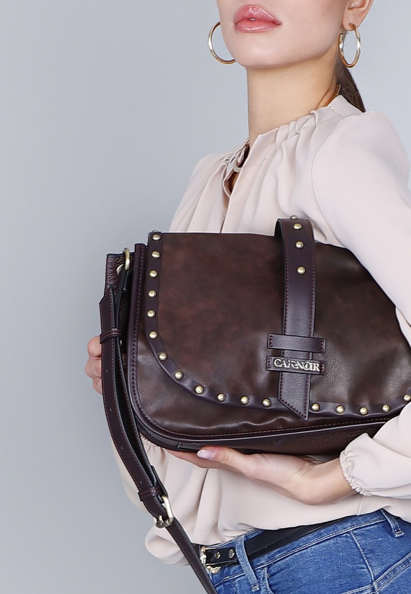 Bruine leren schoudertas met gestikte accenten, verstelbare schouderband en klepsluiting. Gladde textuur met een gestructureerd ontwerp en merklogo.