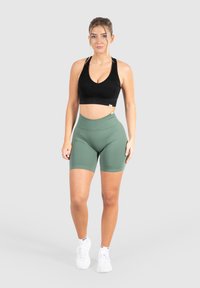 Reggiseno sportivo nero a coste con scollatura a V, abbinato a pantaloni corti verdi a vita alta. Il modello indossa anche sneakers bianche da atletica. Texture liscia.