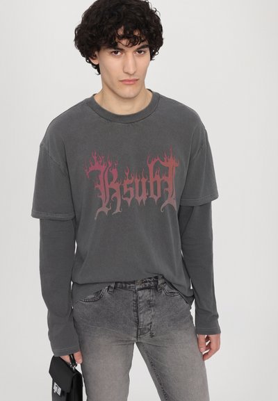 T-shirt gris surdimensionné avec un graphisme "Ksubl" en style flamme rouge, superposé sur une chemise à manches longues grise, accompagné de jeans gris.