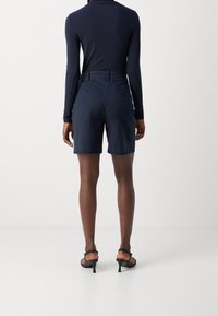 Laivastonsiniset shortsit, joiden pinta on sileä ja vyötärö on istuva, nähtynä takaa. Yhdistetty pitkähiharaiseen paitaan ja mustiin korkokenkiin.