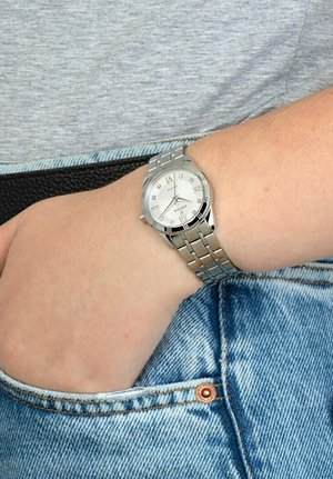 Orologio da polso in metallo argento con quadrante bianco, con indicatori delle ore argentati e bracciale a maglie. Indossato al polso di una persona, esibito su una superficie in denim.