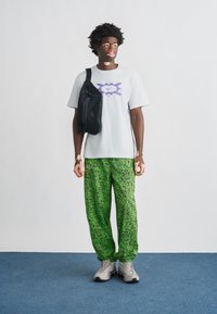 Ανοιχτό γκρι μπλουζάκι με μωβ γραφικό, μαύρη τσάντα χιαστί, πράσινο παντελόνι με σχέδια και γκρι αθλητικά παπούτσια. Απλή και άνετη εφαρμογή.