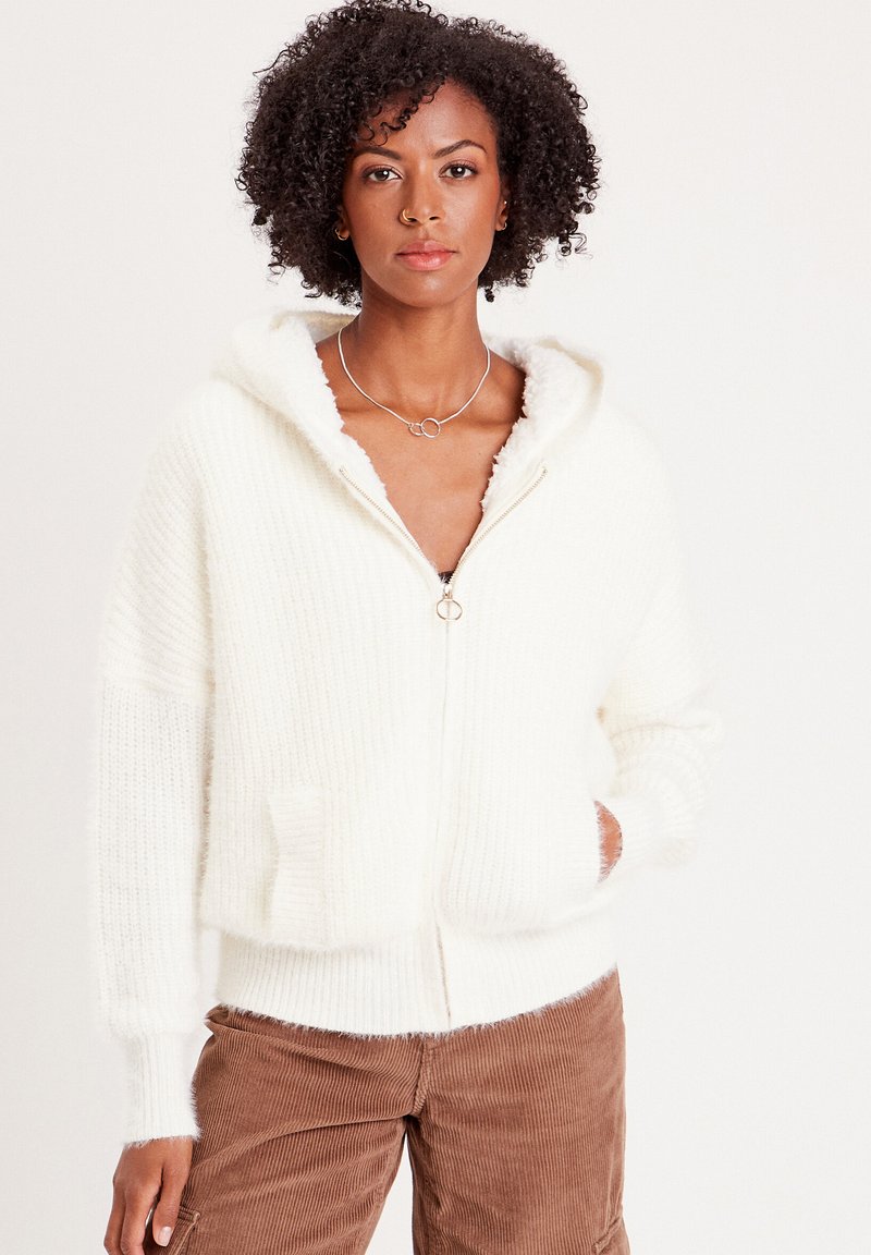 Weißer Zip-Hoodie aus weichem, flauschigem Stoff mit geripptem Textil. Verfügt über eine Kapuze und zwei Fronttaschen. Kombiniert mit braunen Cordhosen.