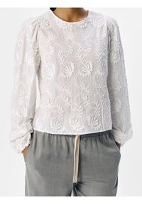 Blouse blanche brodée de motifs floraux, col rond et manches longues bouffantes. Associée à un pantalon gris à cordon de serrage.