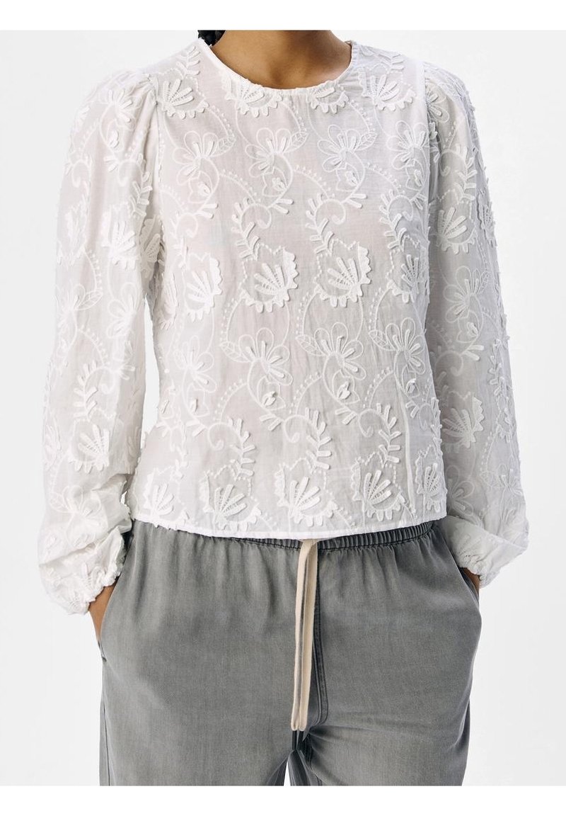 Blouse blanche brodée de motifs floraux, col rond et manches longues bouffantes. Associée à un pantalon gris à cordon de serrage.