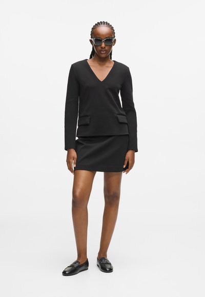 Femme avec des cheveux tressés portant un haut noir à col en V, une mini-jupe noire, des mocassins noirs cloutés et de grandes lunettes de soleil, se tenant sur un fond blanc.