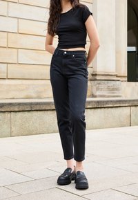 Femme portant un crop top noir, un jean noir taille haute et des mocassins noirs épais, se tenant sur un pavé en pierre extérieur.