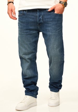 Man wearing dark blue jeans, white sneakers, a white shirt, and a gold watch with a black leather band.