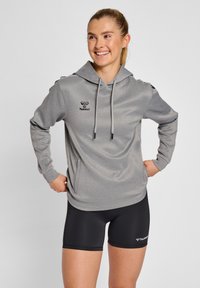Haut à capuche gris à manches longues avec une capuche à cordon de serrage, logo sur la poitrine et accents aux épaules. Associé à un short de sport noir.