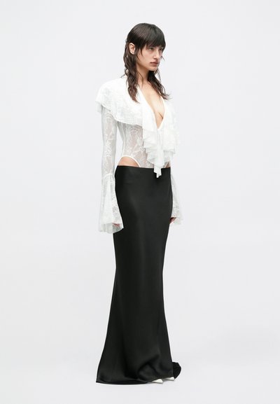 Γυναίκα που φοράει μακρύ μαύρο φούστα και διάφανο λευκό δαντελένιο τοπ με βαθύ V λαιμόκοψη και βολάν, στέκεται μπροστά σε ένα απλό λευκό φόντο.