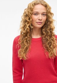 Punainen kuvioitu villapaita pyöreällä pääntiellä ja pitkillä hihoilla, jossa korotettu silmukkakuvio. Mallilla on aaltoilevat, olkapäille ulottuvat hiukset.