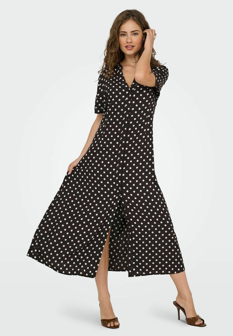 Robe noire à pois blancs, à manches courtes, avec un devant à boutons et une fente haute. Fabriquée en tissu doux, avec des pois blancs répartis sur l'ensemble.