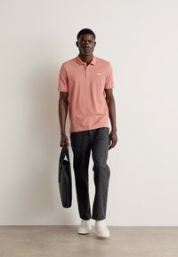Chemise polo rose, jeans foncés, tenant un sac noir, debout sur un fond neutre. Le look présente des manches courtes et un col.