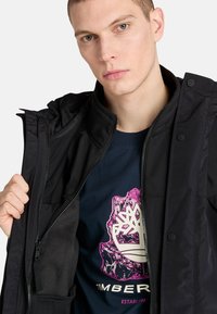 Musta tuulenpitävä takki, jossa on vetoketjukiinnitys ja nepparit, kerrostettuna laivastonsinisen T-paidan päälle, jossa on vaaleanpunainen ja valkoinen puun logo.