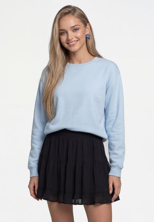 Lachende vrouw met lang blond haar draagt een lichtblauwe trui, een zwarte geplooide rok en blauwe stervormige oorbellen tegen een witte achtergrond.