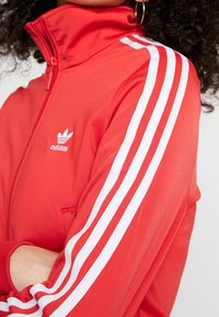 Červená zipová bunda s vysokým límcem, bílými třemi pruhy na rukávech a malým bílým logem Adidas na hrudi. Hladká, pružná tkanina.