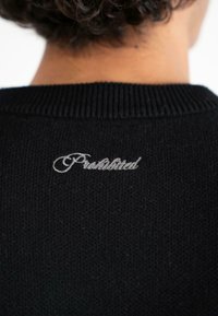 Vista posteriore di una persona che indossa un maglione di lana nera con ricamo bianco in corsivo che legge "Proibito" sulla parte superiore della schiena.