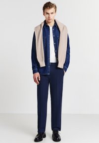 Camicia di jeans blu scuro, t-shirt bianca a girocollo, pantaloni blu navy, maglione beige lavorato a maglia drappeggiato sulle spalle, scarpe nere con lacci.