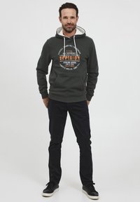 Sudadera verde oscuro con cordones blancos, que presenta un gran logo impreso en naranja y negro. Combinada con pantalones oscuros y zapatillas.