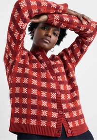 Žena oblečená v červeno-bielo-tmavočervenom vzorovanom pletenom kardigáne s gombíkmi, zdvíha ruky nad hlavu pred jednoduchým pozadím.