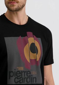 Mann trägt ein schwarzes T-Shirt mit buntem abstraktem Grafikdesign und dem Text "Pierre Cardin 1954" auf der Vorderseite.