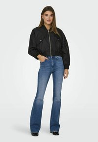 Veste bomber noire courte avec fermeture éclair argentée et poches à pression, associée à un jean en denim bleu évasé et des chaussures noires. Texture lisse.