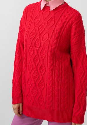Roter, grob gestrickter Pullover mit Rauten- und Zopfmustern, getragen über einem hellrosa Hemd mit Kragen und violetten Hosen.